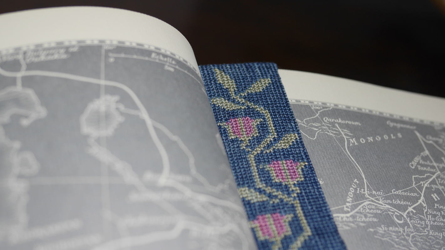 Un marque page brodé dans un livre ouvert. La broderie est une frise fleurie sur fond bleu, avec des roses à peine écloses.