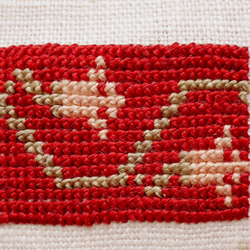 Une frise brodée sur fond rouge brodée au point de croix natté, une tige qui forme des S avec des fleurs rose pâles de chaque côté.