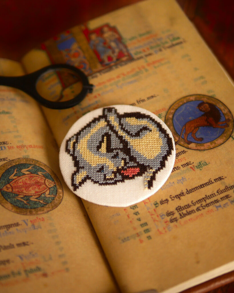 Petit médaillon brodé au point de croix : un chat qui se lave le cul dans le style des enluminures médiévales. Le médaillon est posé sur la reproduction d'un manuscrit enluminé avec la représentation du lion (le signe du zodiaque).