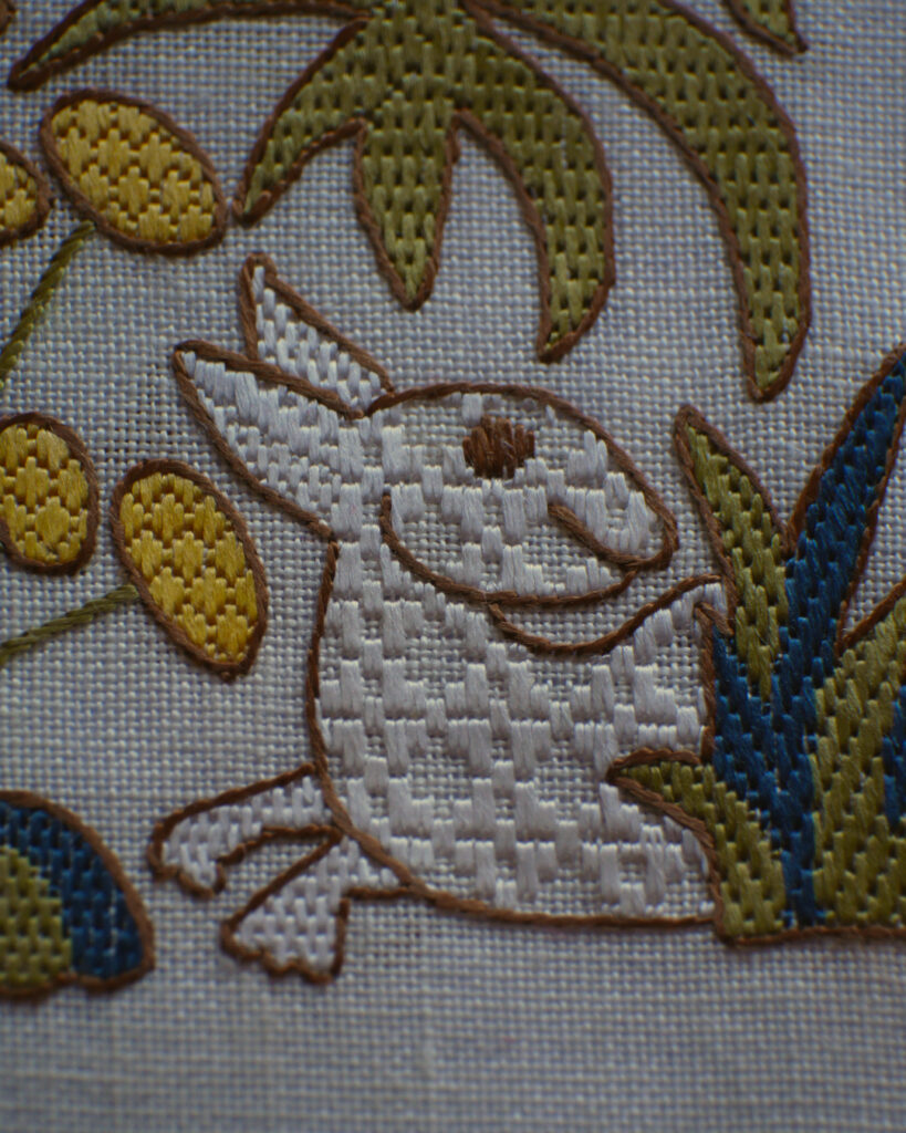 Détail d'une broderie dans le style médiéval à points comptés. Un petit lapin sort d'un buisson d'herbe, on ne voit pas l'arrière de son corps, il a la tête tournée vers l'arrière. Il est brodé au fil blanc sur toile blanche avec des points réguliers qui forment une texture géométrique laissant apparaître la toile de fond. Son œil et le contour sont brodés en marron. Autour de lui, on devine des fleurs et végétaux brodés en fils colorés toujours avec des points géométriques.