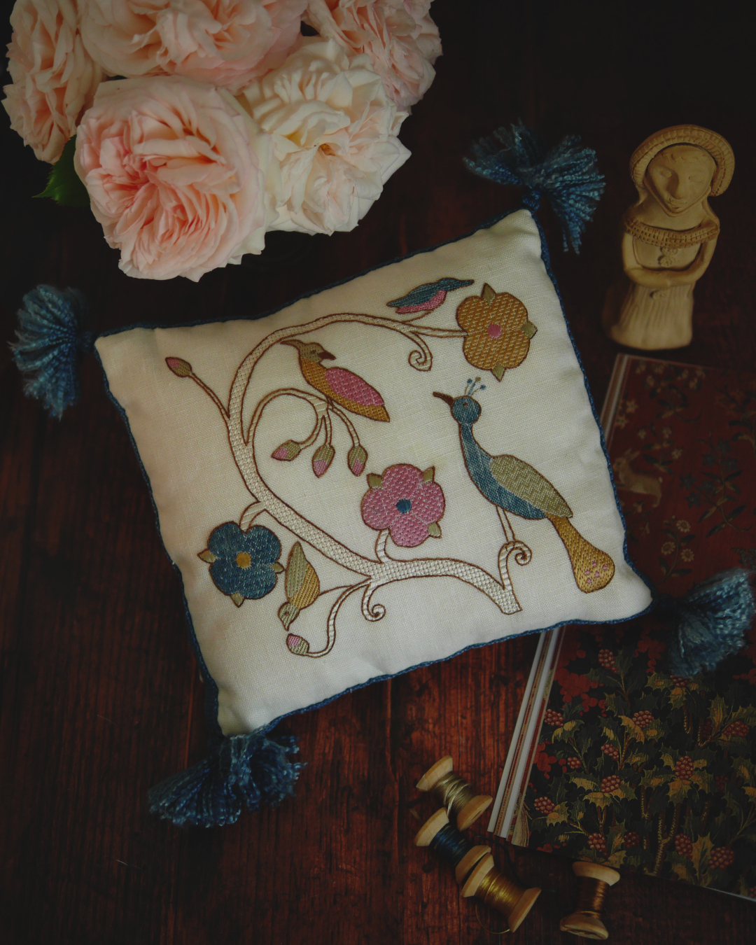 Coussin brodé dans un style pastel et médiéval : une branche de rosier avec des roses stylisées et des bourgeons où sont posés 4 oiseaux : un paon, une huppe, un martin pécheur et un rossignol. Avec des points de broderies réguliers qui forment des textures différentes sur chaque oiseaux, chaque fleur. Il y a également un bouquet de roses blanches, une poupée médiévale en céramique et un livre ouvert.