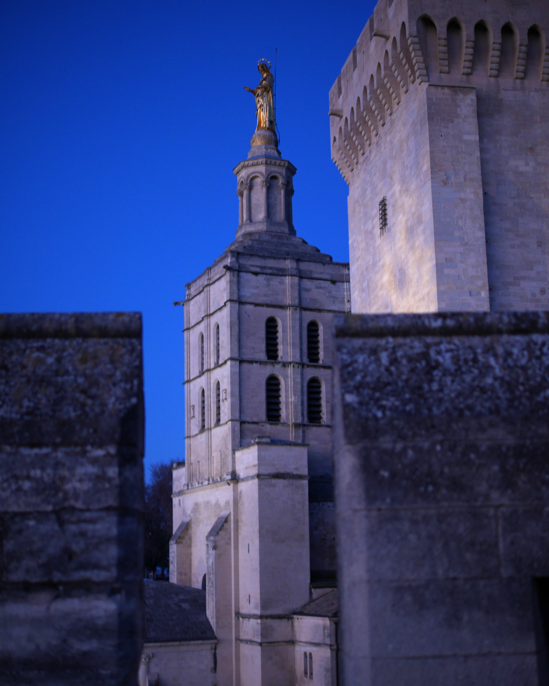 Photo de l'extérieur du palais des papes à Avignon. Les remparts en pierre blanche et une tour avec une statue de vierge dorée au sommet.