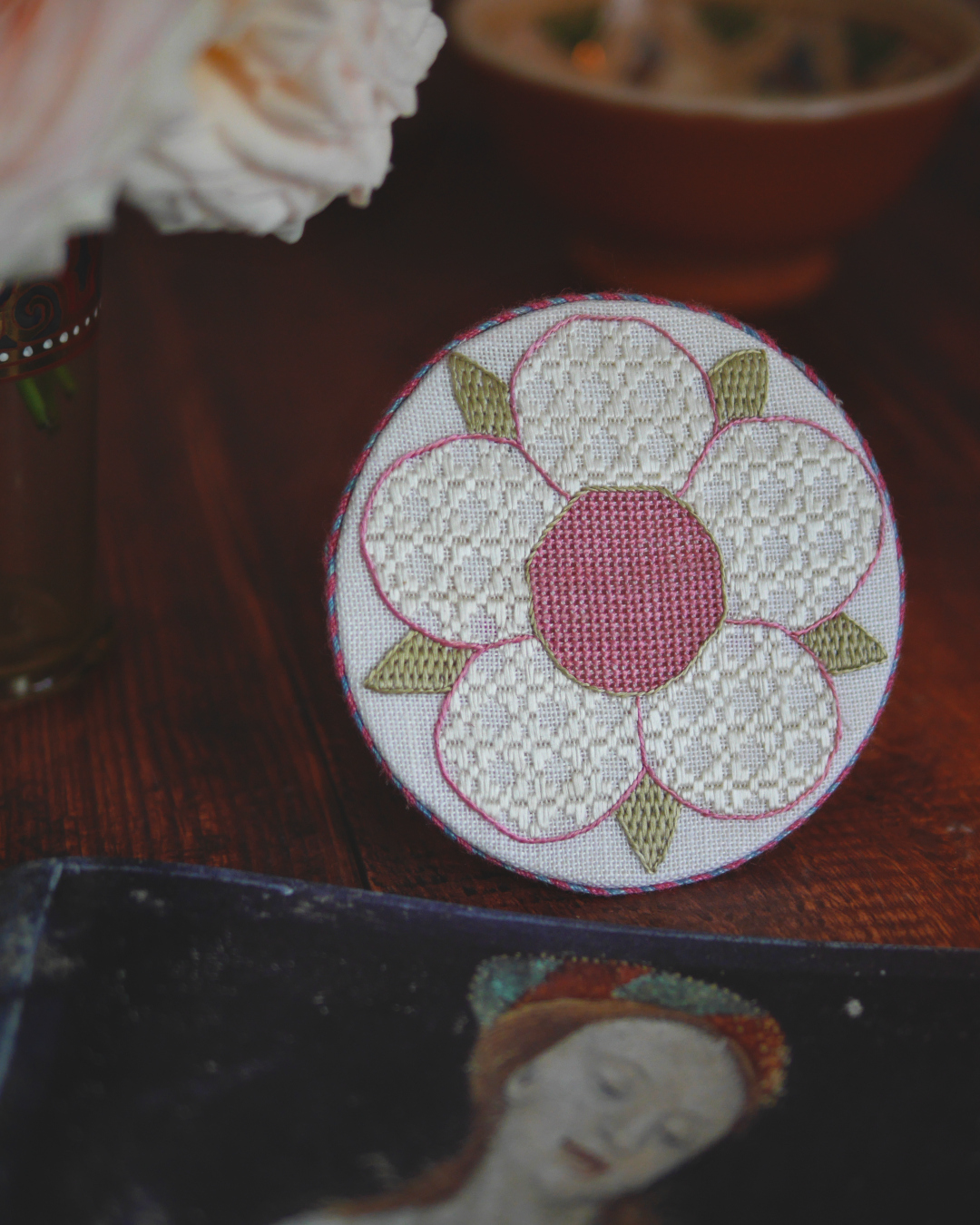 Médaillon brodé d'une rose médiévale simplifiée : 5 pétales blancs, un cœur rose pastel et des sépales vert pastel. Tous les remplissages sont réalisés avec des points géométriques qui créent des textures différentes. Devant un livre ouvert.