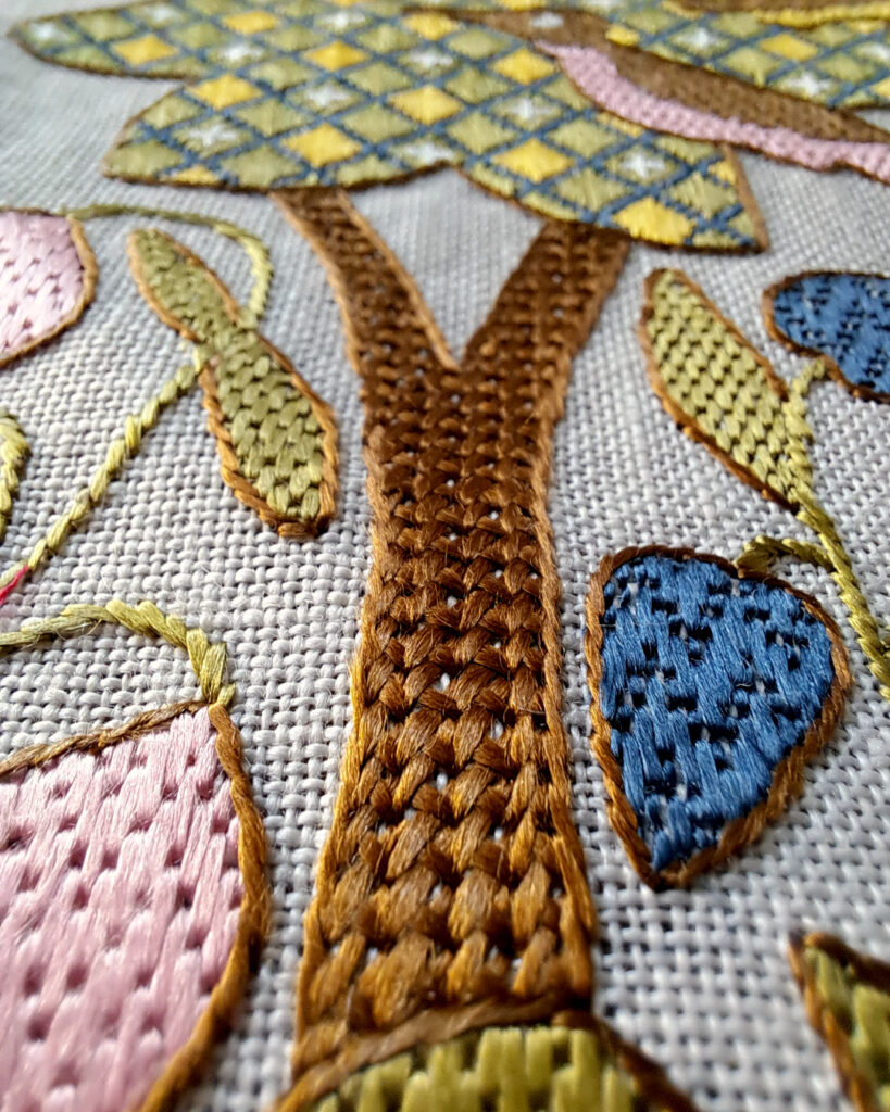 Détail d'une broderie dans le style médiéval.
Un tronc brodé en marron au point de croix natté en lignes verticales.
Autour des feuilles et des fleurs roses, bleues et vertes.