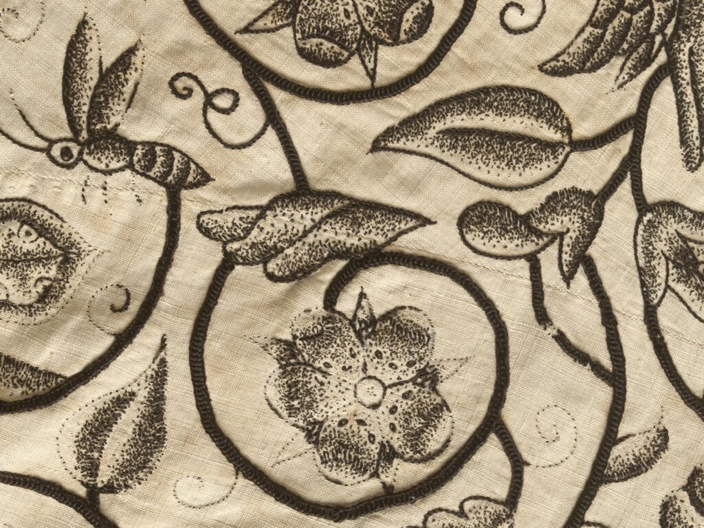 Détail d'une broderie au fil noir sur toile écrue. La broderie est un décor mélangeant végétaux et animaux dans le style des gravures. Les tiges qui forment un décor sont brodées avec un point plus épais.