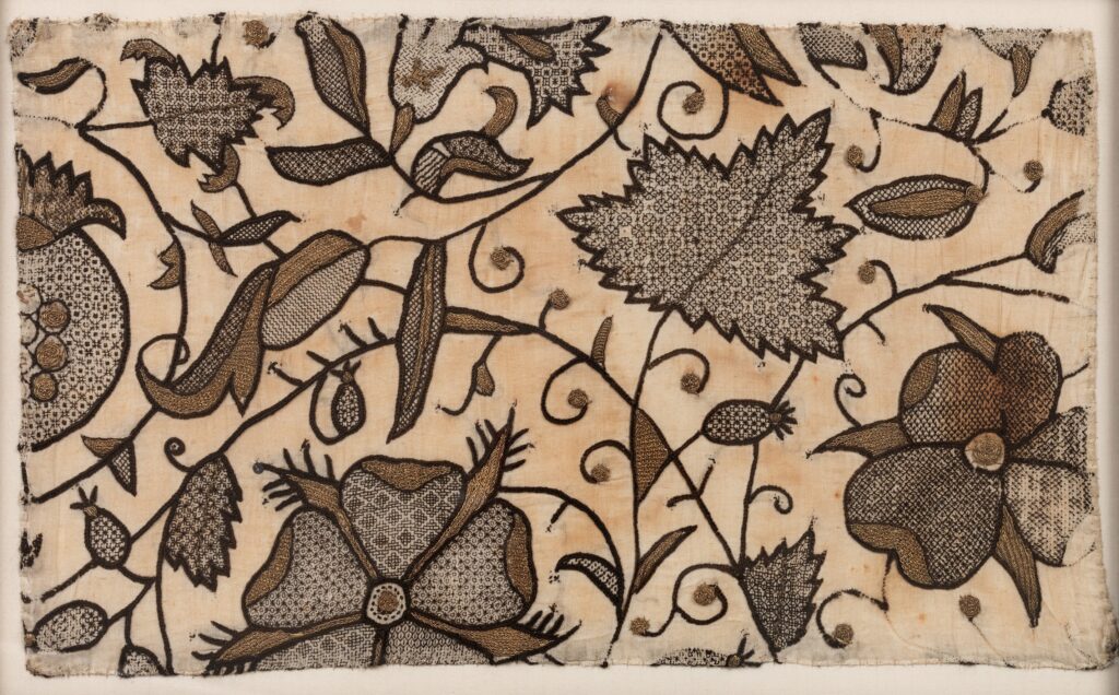 Broderie sur toile écrue au fil noir et au fil doré. C'est un décors végétal avec des points linéaires pour les contours et les tiges et des motifs géométriques au fil noir pour le remplissage. Des détails sont brodés au fil d'or avec des points tressés