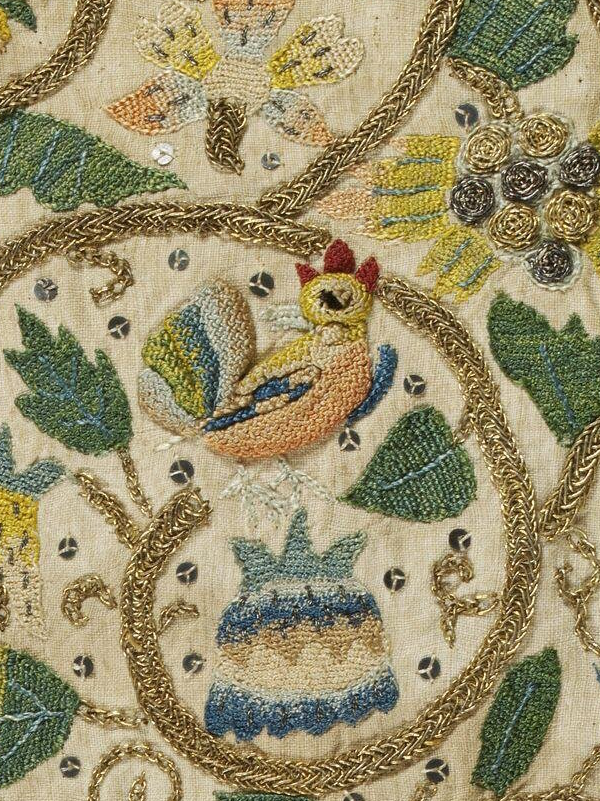 Détail d'une broderie sur toile écrue avec un décor végétal et un coq coloré au centre. La broderie est réalisée avec des fils colorés et les tiges sont brodées au fil d'or avec un point tressé.