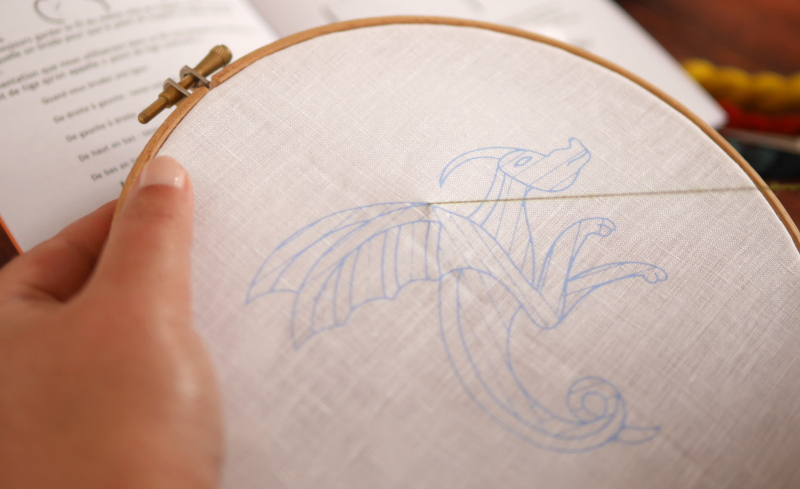 Un tambour de broderie tenu en main. Sur la toile blanche, un dragon est imprimé avec des lignes espacées sont imprimées.